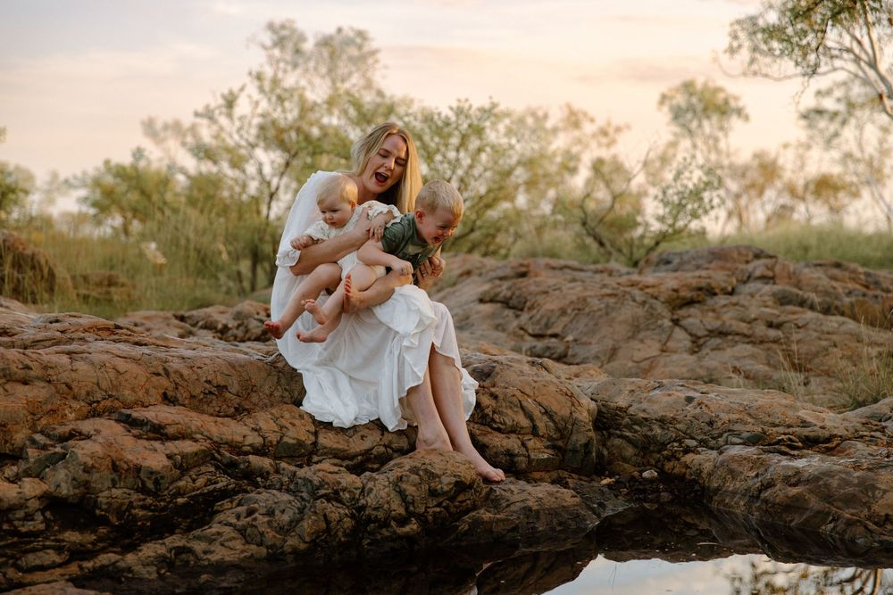 A figure in a flowing white dress sits on rocky terrain at sunset with trees silhouetted in the background.