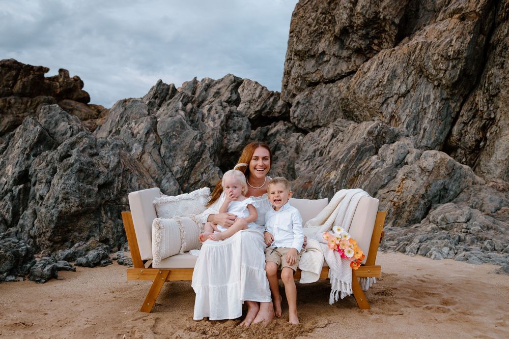 A beige vintage couch sits on a sandy beach against dramatic rocky cliffs during a sunset photoshoot.
