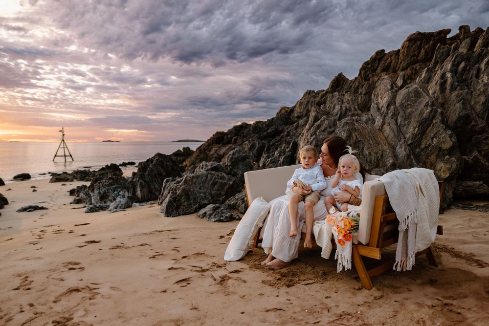 A family relaxes together on a white couch at the beach during sunset with rocky cliffs and ocean in the background.