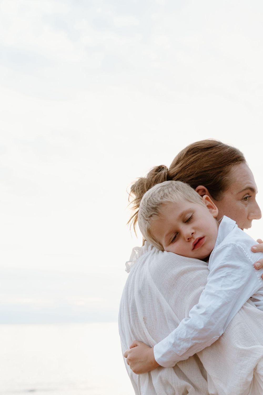 A tender moment captured as a sleeping child in white rests peacefully against someone's shoulder outdoors.