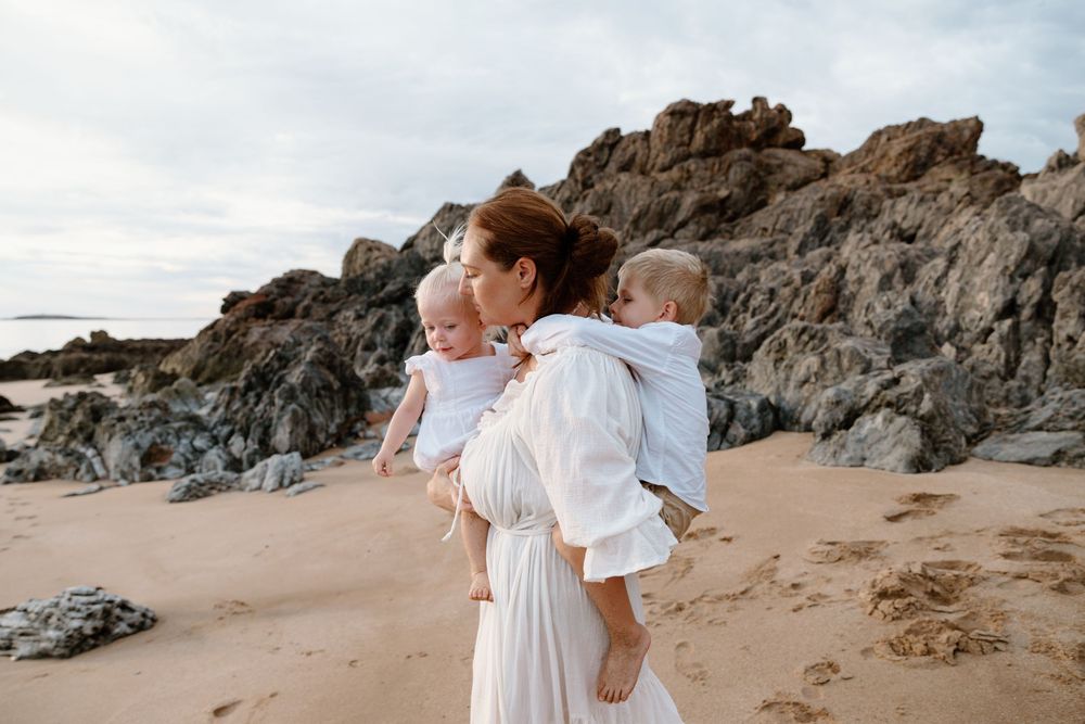 A person in white holding two small children on a scenic rocky beach at sunset.