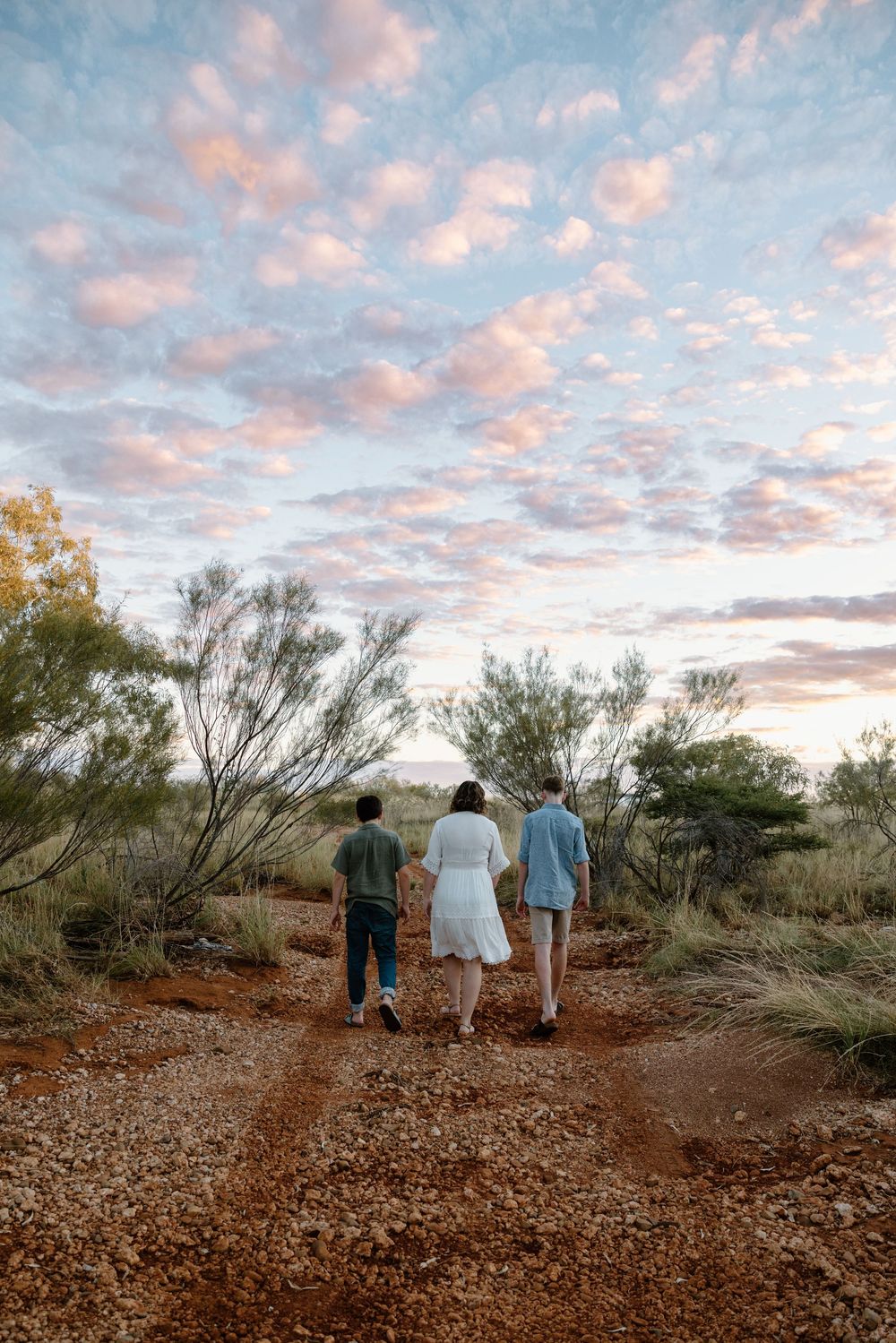 Three people walk along a rocky desert trail at sunset with pink clouds overhead.