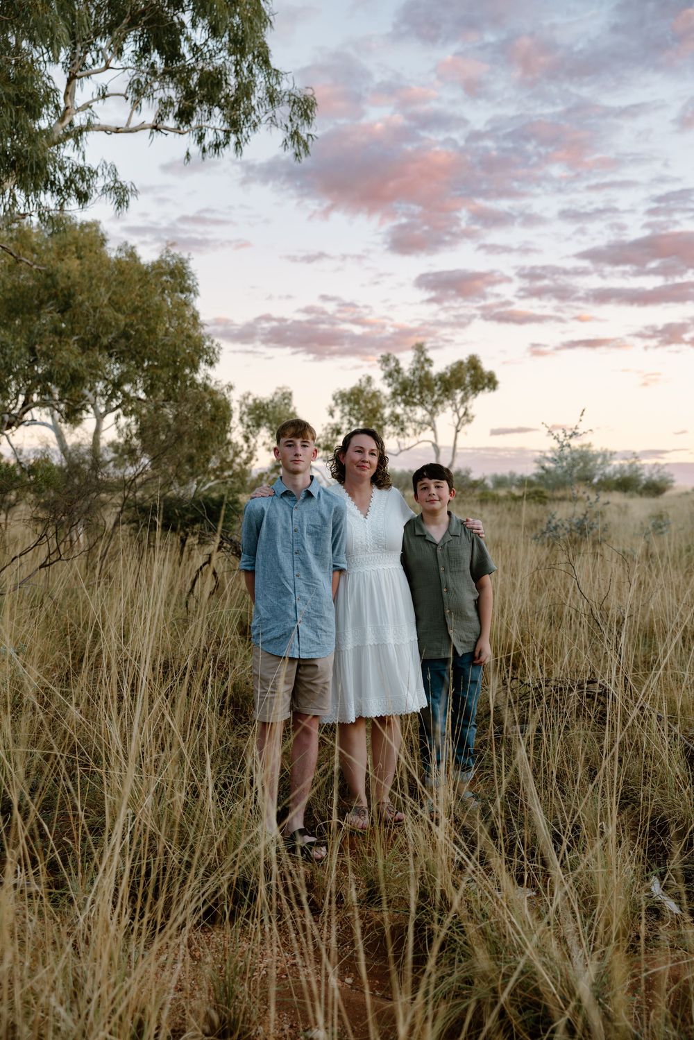 Three people stand in golden grass field at sunset with eucalyptus trees and pink clouds in the background.