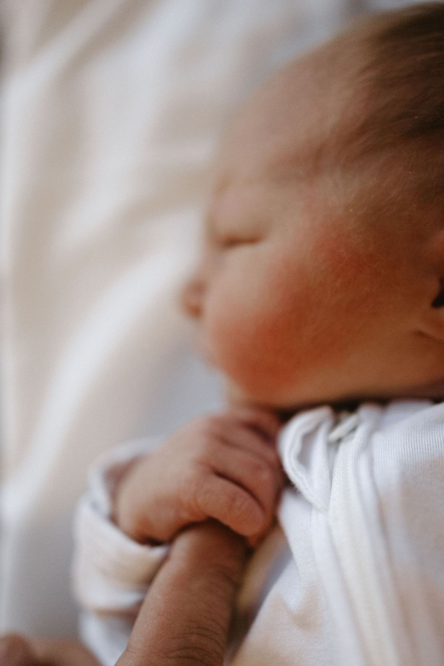 A newborn infant sleeps peacefully in soft white clothing while grasping their tiny hand against their chest.
