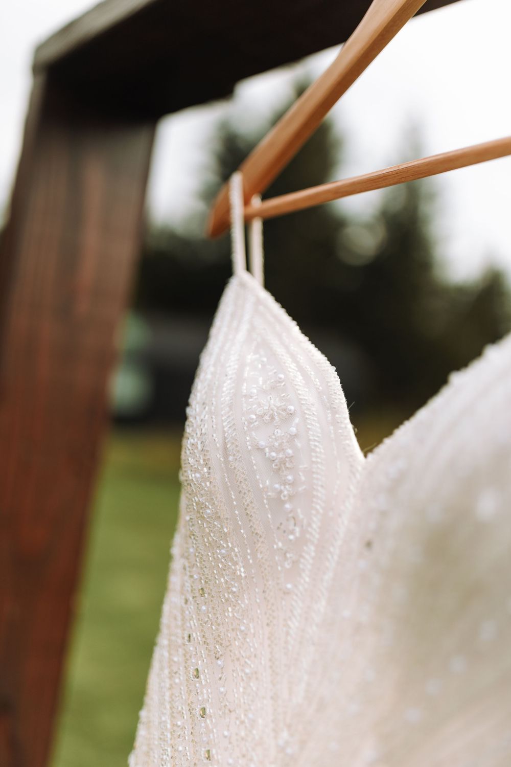 Close-up of a white lace wedding dress detail with water droplets hanging on wooden hangers outdoors.