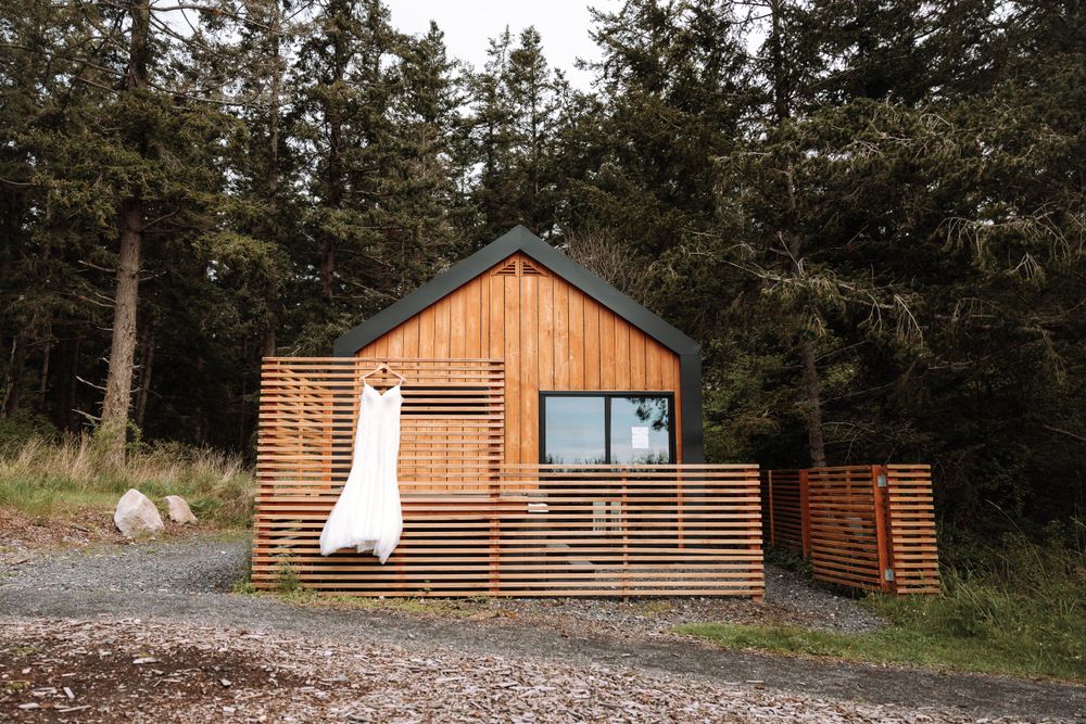 A modern wooden cabin with horizontal slat siding and a white dress hanging on the deck in a forest setting.