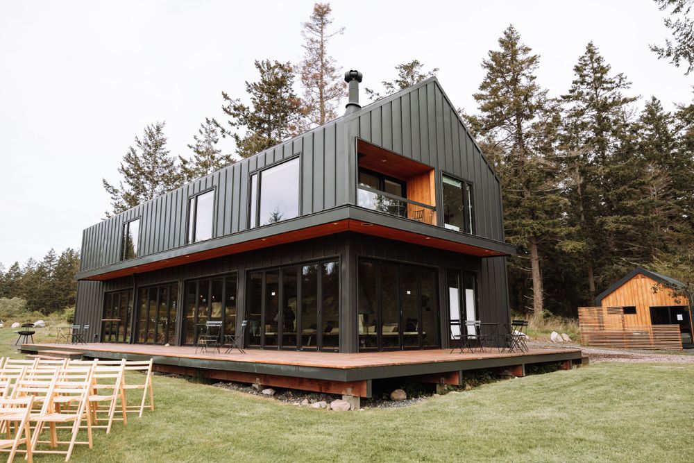 Modern dark gray metal-clad house with large glass windows and wooden deck surrounded by evergreen trees on grassy lawn.