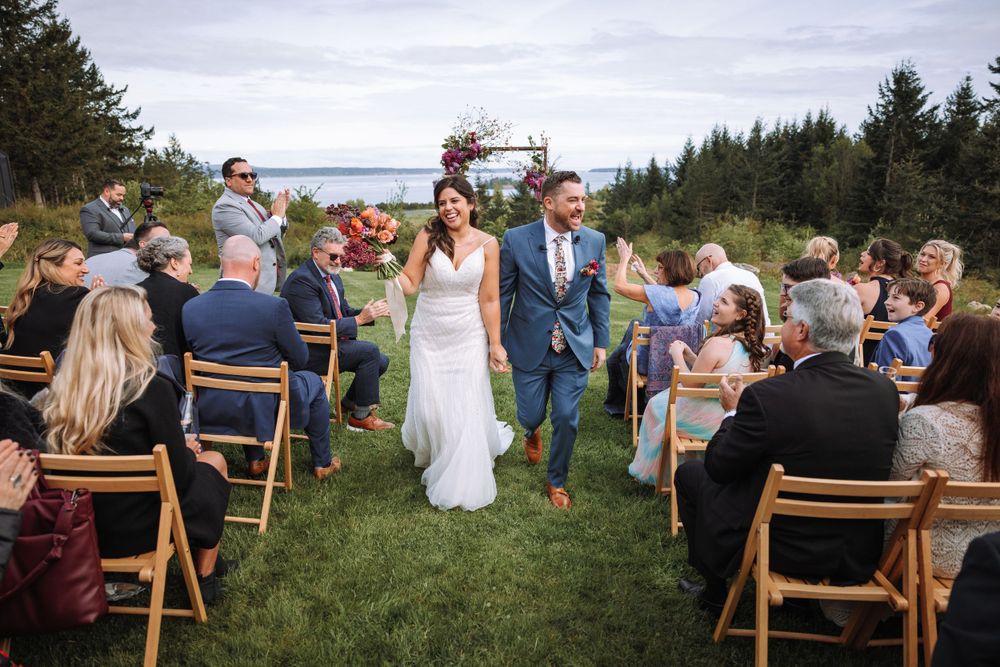 A wedding ceremony takes place on a scenic hilltop overlooking water, with guests seated on wooden chairs among evergreen trees.