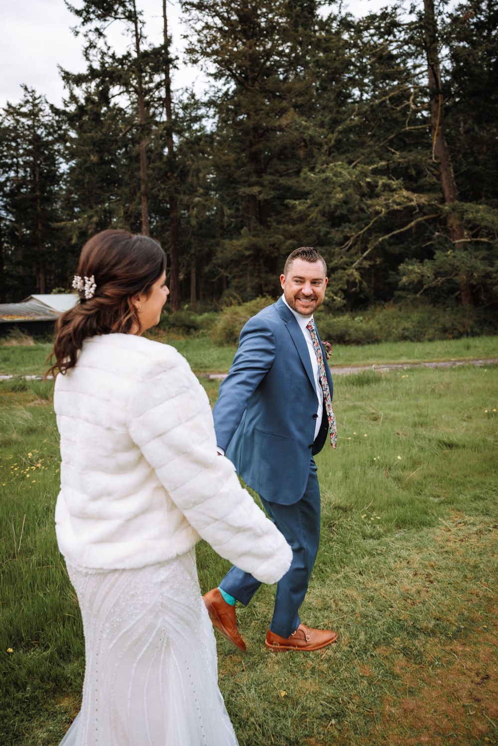 A couple in wedding attire share a playful moment in a woodland meadow with evergreen trees in the background.
