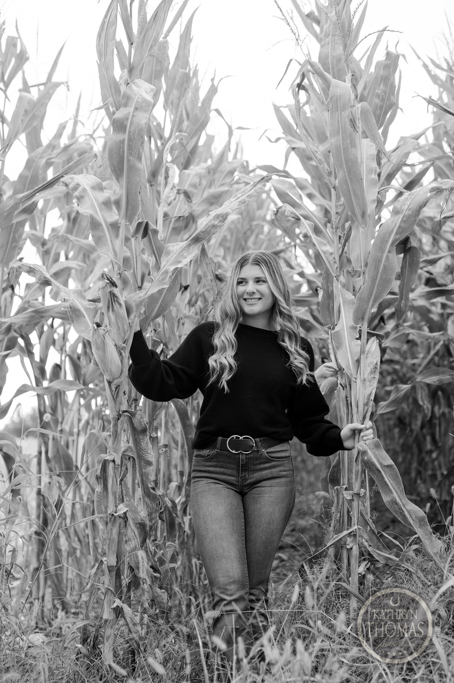Black and white photo of someone posing between tall corn stalks in casual fall attire.