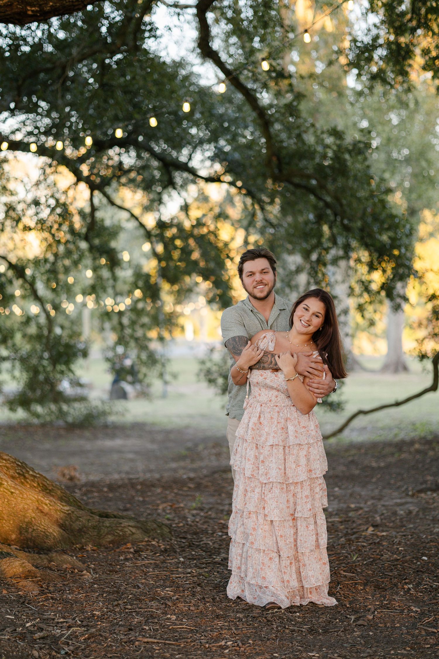 Couple embracing outdoors under green trees at golden hour, woman in floral dress, man in green shirt, smiling.