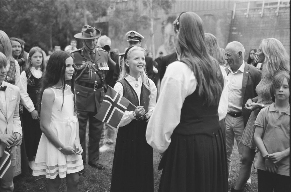 Children in traditional Norwegian bunad costumes holding flags at a National Day celebration outdoors.