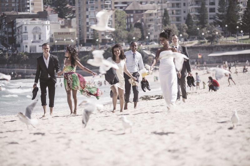 A wedding party walks together along a sunny beach with buildings in the background.