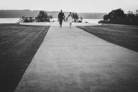 Two silhouettes walk along a paved pathway beside water at dusk.