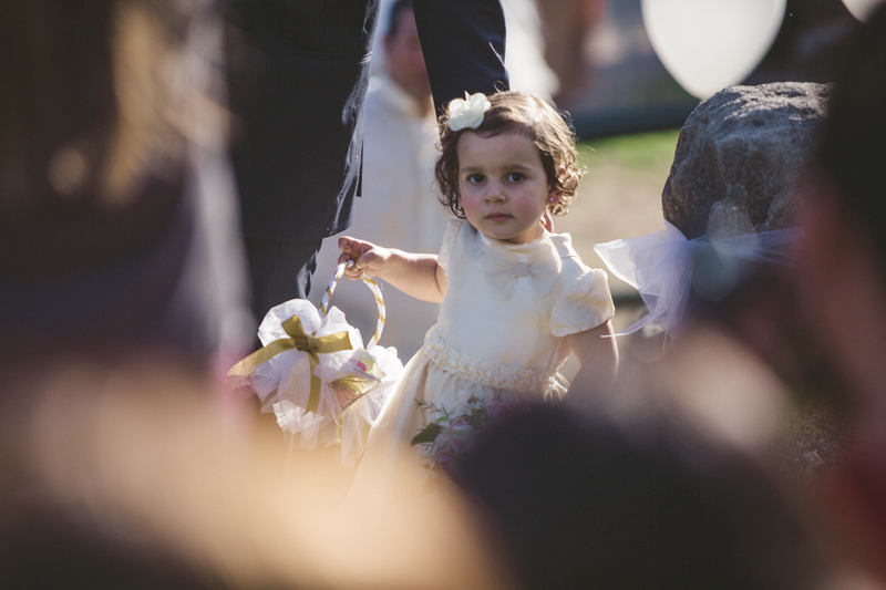 A small child in white dress carries flowers during an outdoor celebration.