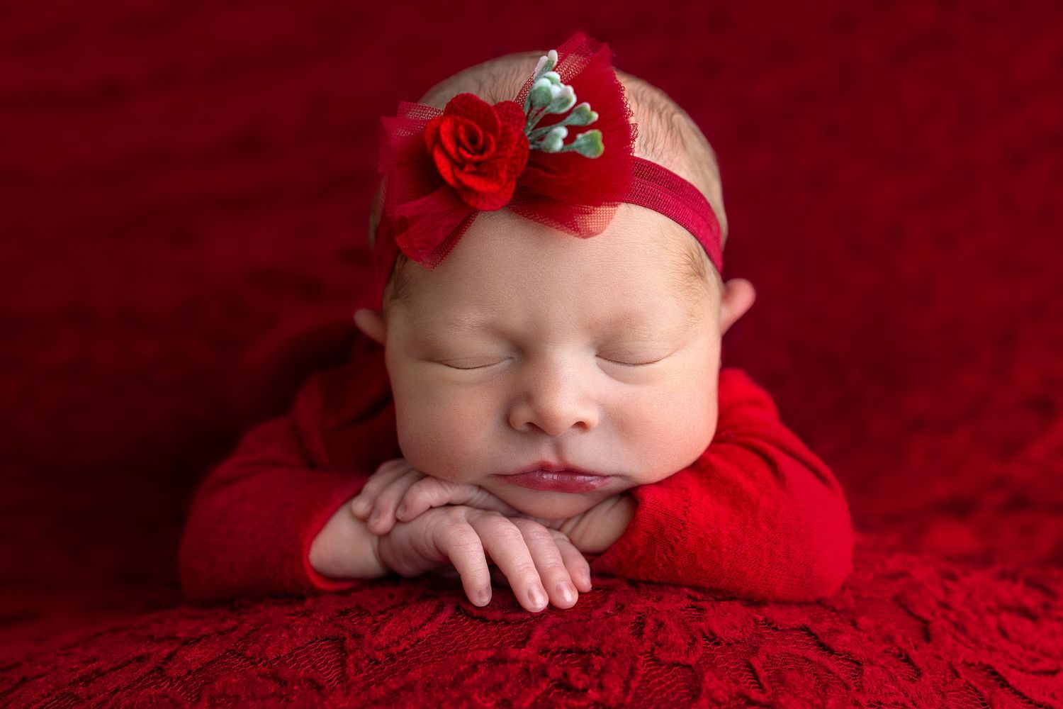 Sleeping newborn in red outfit with floral headband resting on soft red textured fabric background.