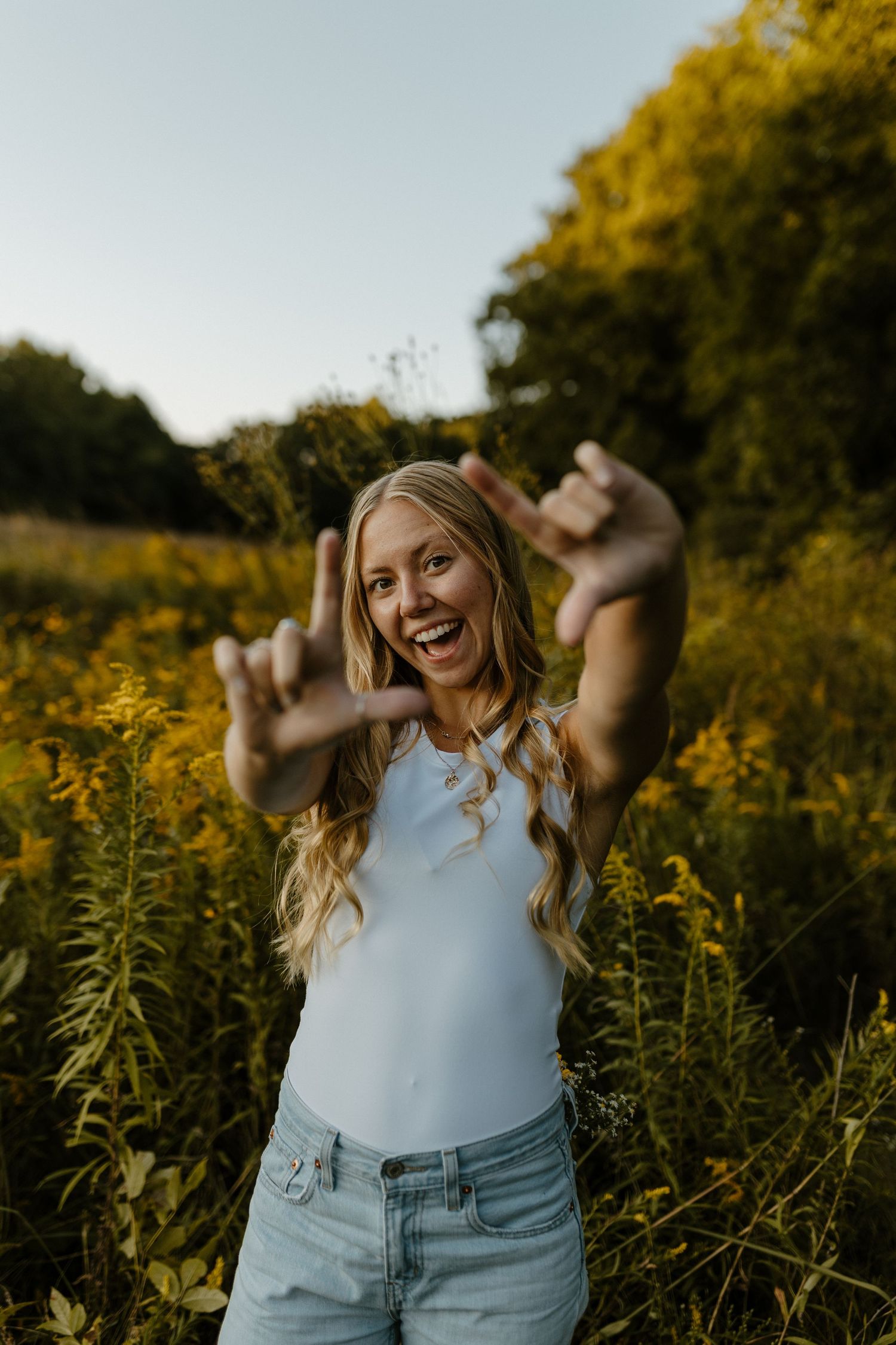 A smiling young woman with long blonde hair stands in a field of yellow flowers, raising her arms in a carefree gesture. Fort Worth Texas