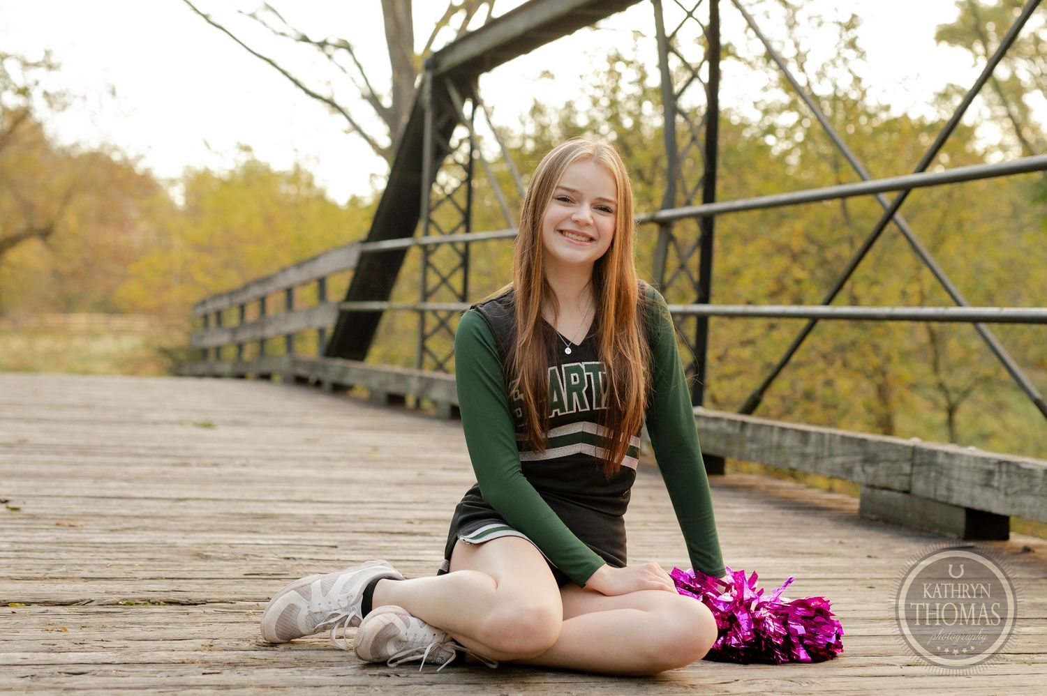 Cheerleader in green and black uniform sits with pom poms on a wooden bridge during autumn.