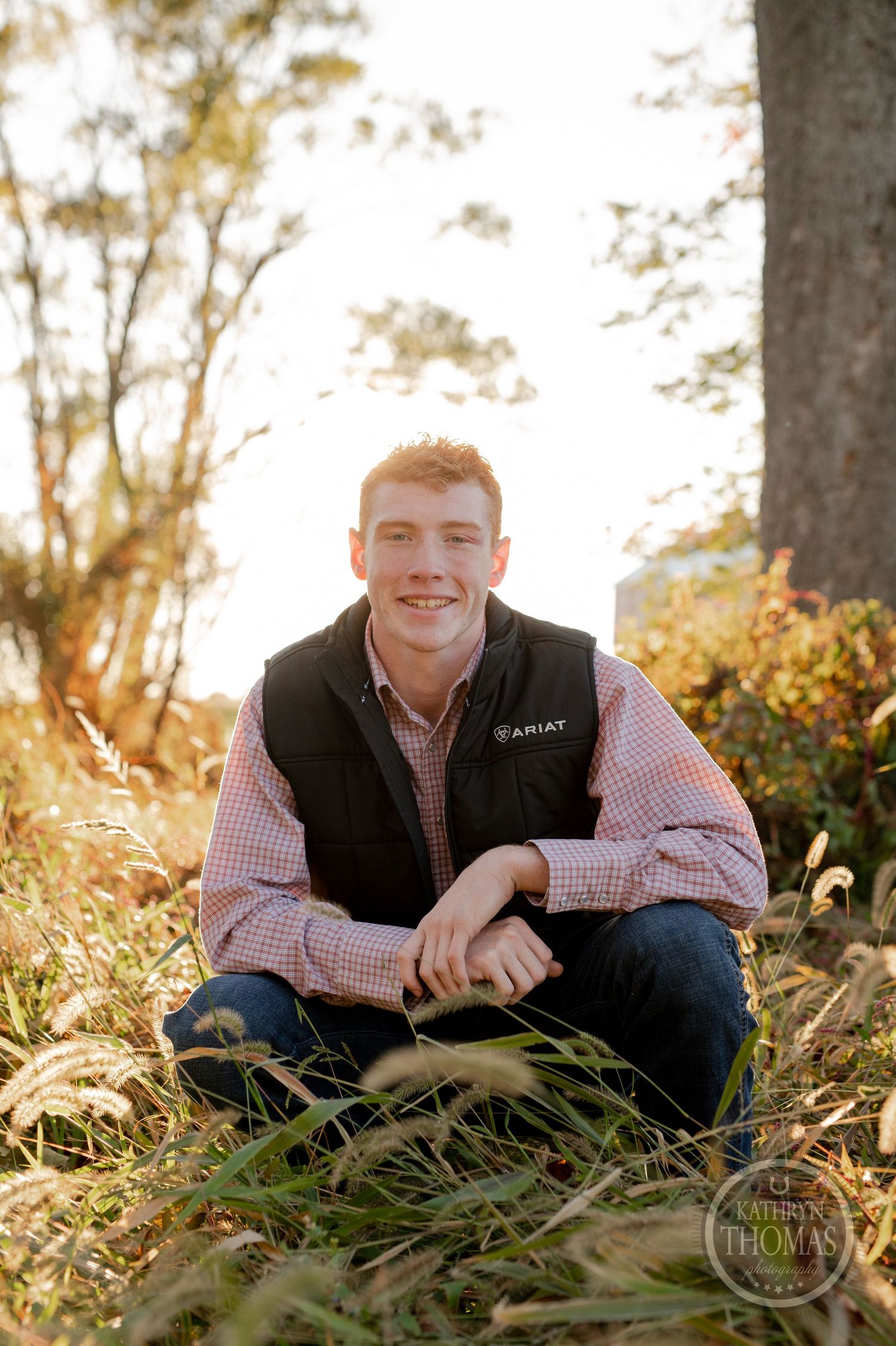 Person sitting in tall grass wearing black vest and plaid shirt with warm sunset lighting.