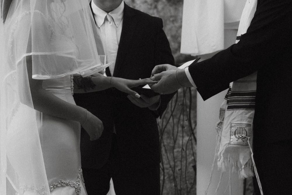 Black and white photo of a wedding ceremony showing hands exchanging rings with officiant and witnesses present. Timeless detail wedding