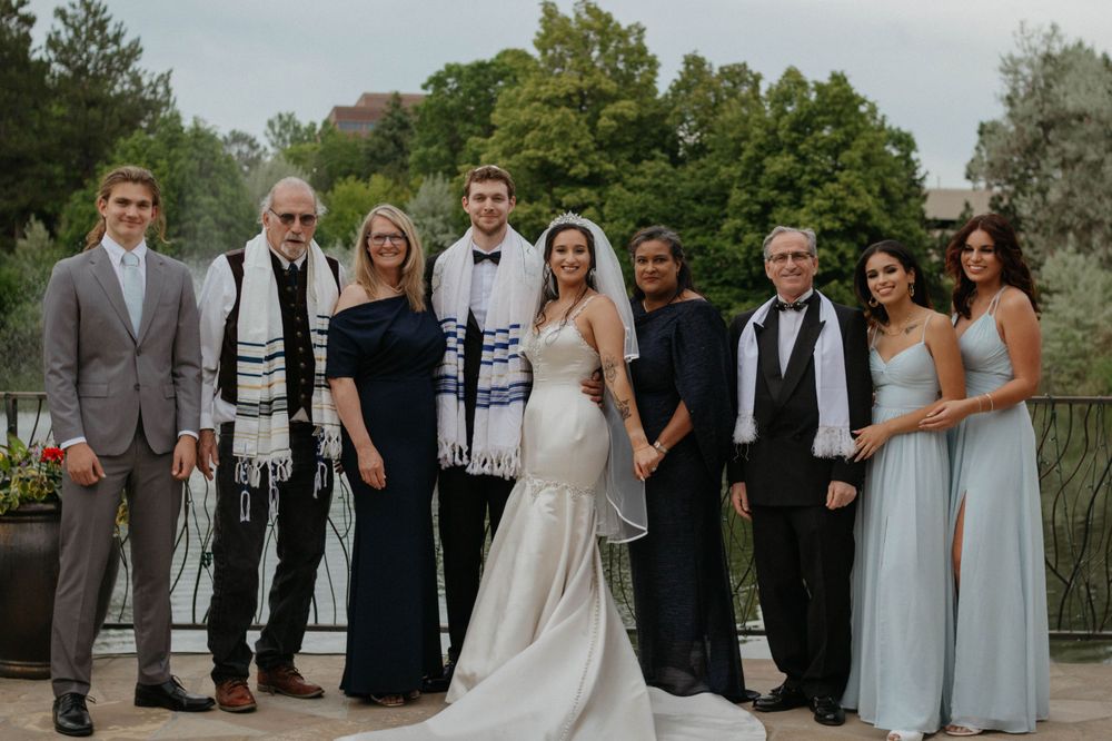 A formal wedding party wearing elegant attire poses together on a stone terrace with lush greenery in the background. Denver, Colorado