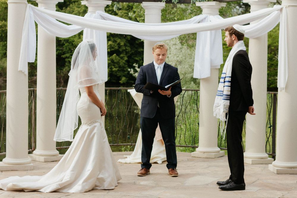 An outdoor wedding ceremony takes place under a white draped gazebo with white columns and green foliage in background in Denver, Colorado