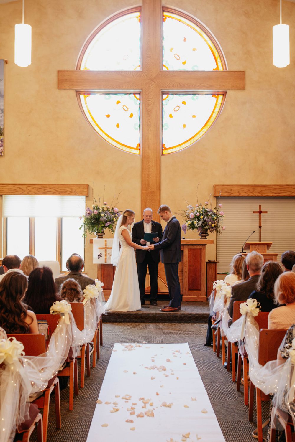 Wedding ceremony taking place in a church sanctuary with a large wooden cross and window. Candid wedding photography in Colorado