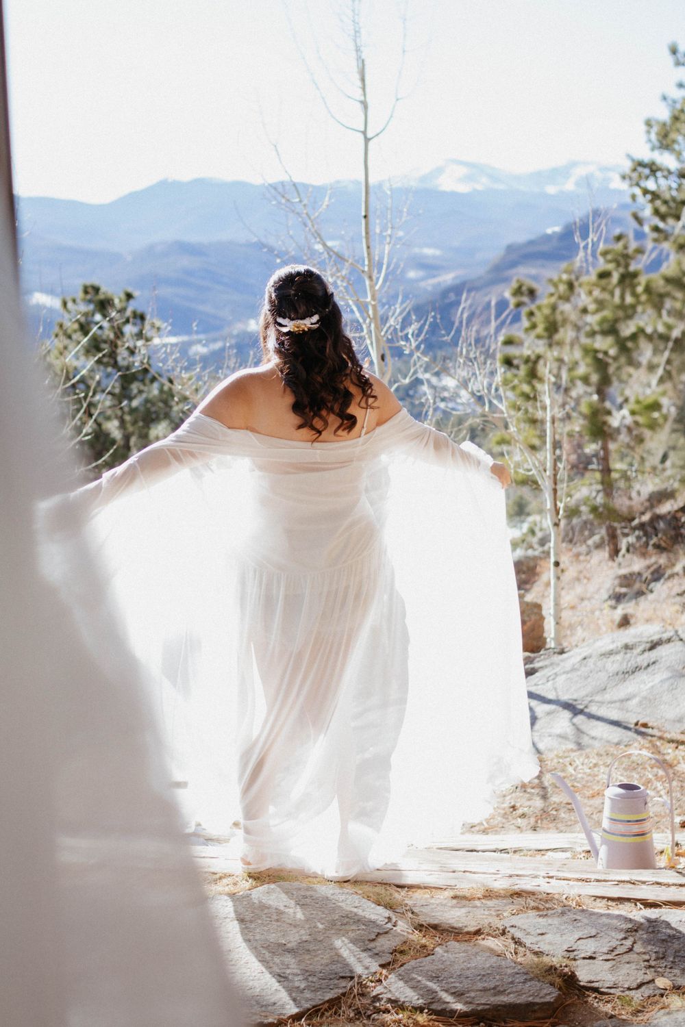 Person in flowing white dress stands on mountain ledge overlooking snowy peaks in romantic outdoor setting in Colorado. Bride getting ready