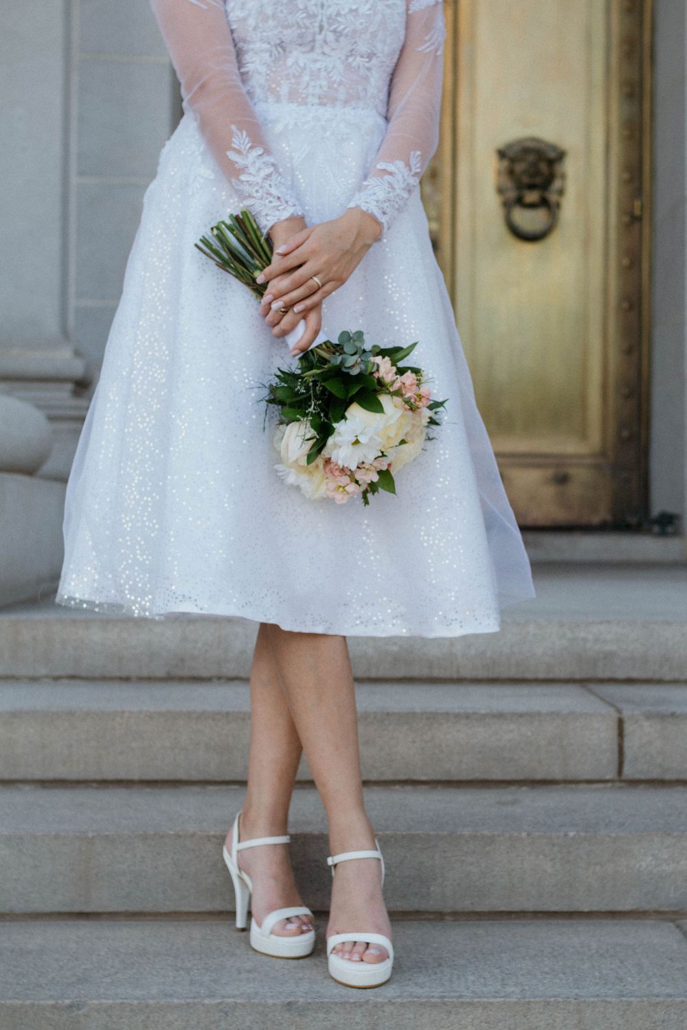 Bride in tea-length lace wedding dress and white sandals holds delicate peach and white bouquet on stone steps.