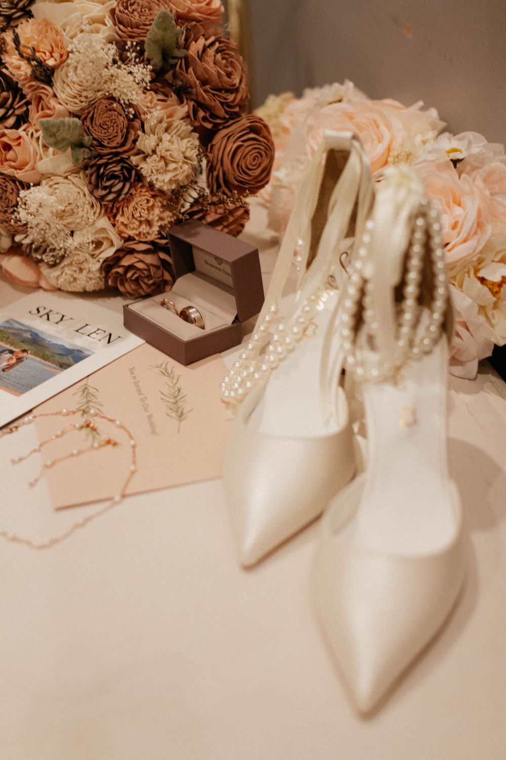 Elegant white bridal shoes with pearls next to dried brown roses and floral arrangements on a light surface. wedding details