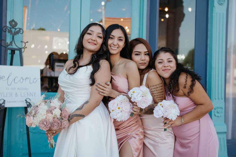 A group in matching pastel dresses poses together with white bouquets outside a turquoise storefront. Bridal party portrait