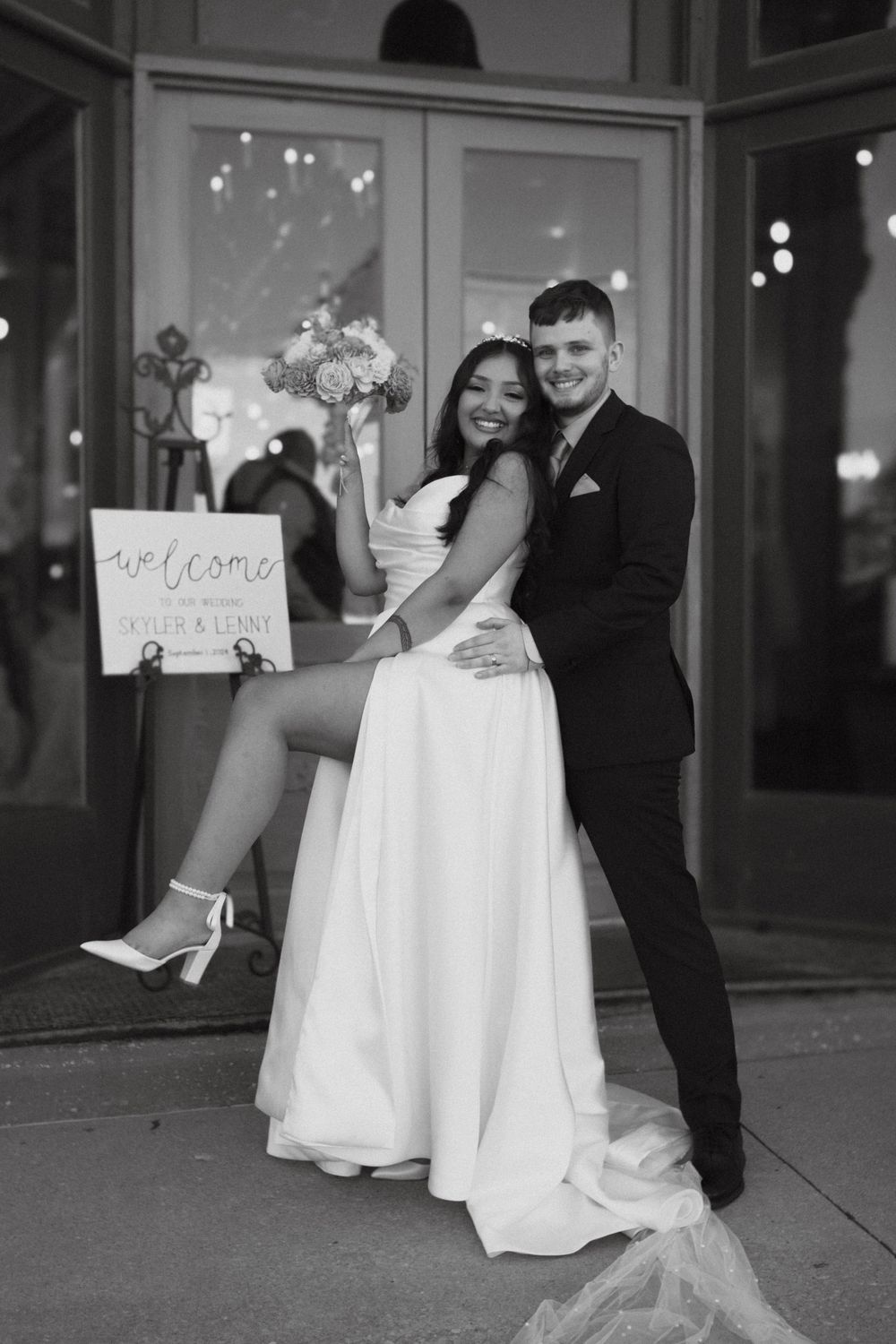 A black and white wedding photo showing a couple embracing outside a venue with a welcome sign and twinkle lights.