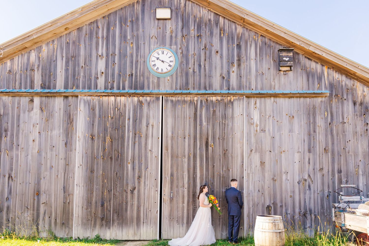 Bride and groom standing in front of weathered barn with rustic wooden siding.