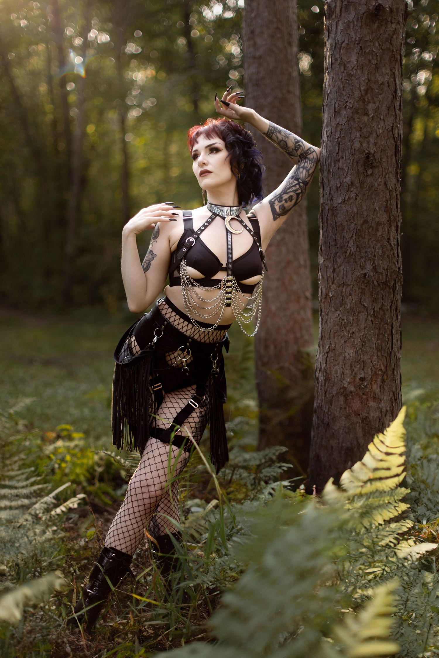 Gothic model in black vintage-inspired burlesque outfit poses dramatically in a forest with ferns and golden sunlight.
