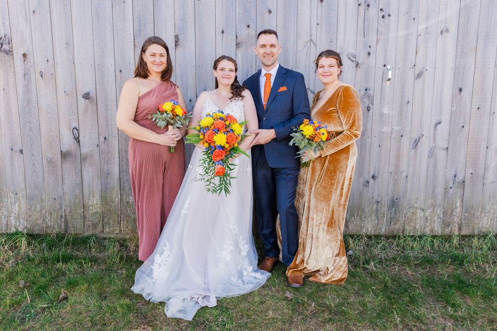 Wedding party of four poses against wooden barn wall with bright autumn bouquet.