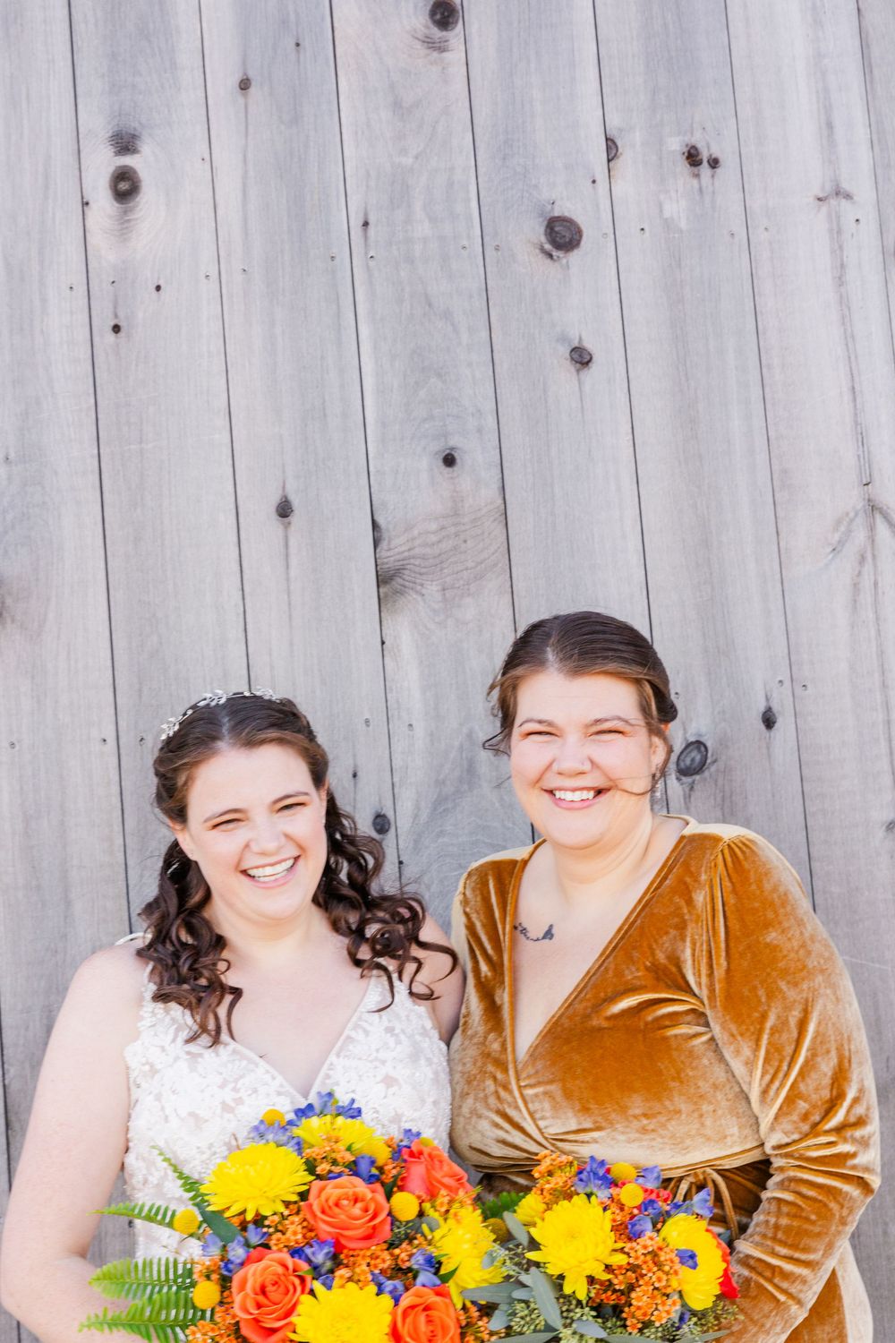 Two women smile brightly against wooden wall, one in wedding dress holding colorful bouquet.
