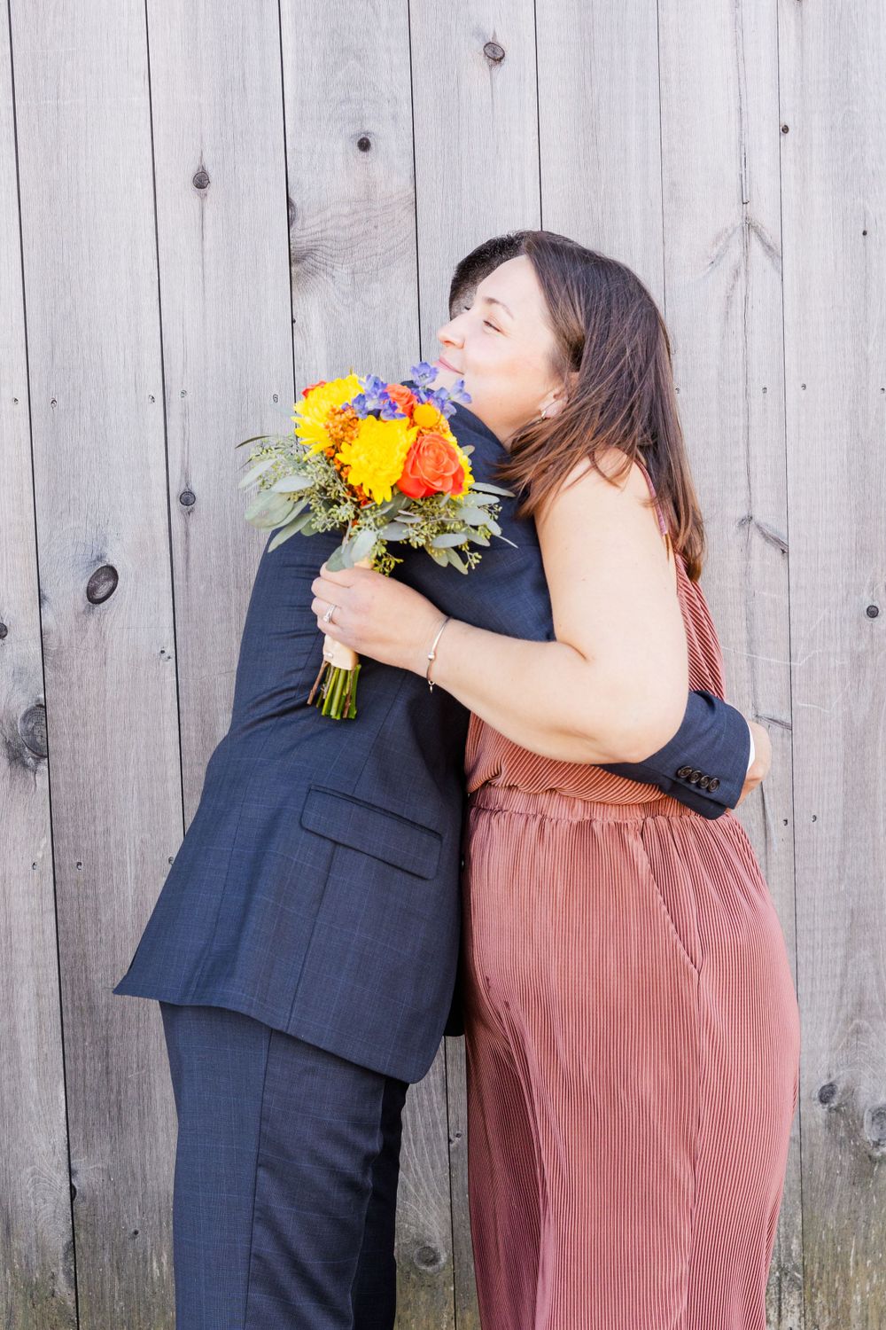 A couple embraces next to a rustic wooden fence while holding a vibrant yellow and orange bouquet.