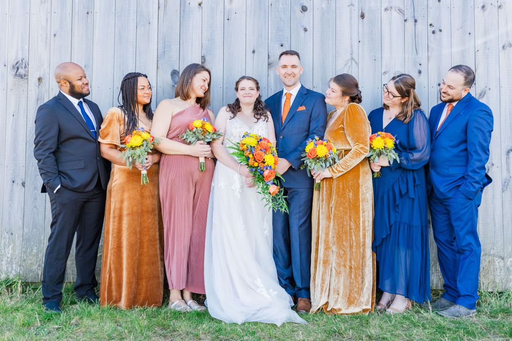 Wedding party in jewel-toned dresses poses against rustic barn wall with bright floral bouquets.