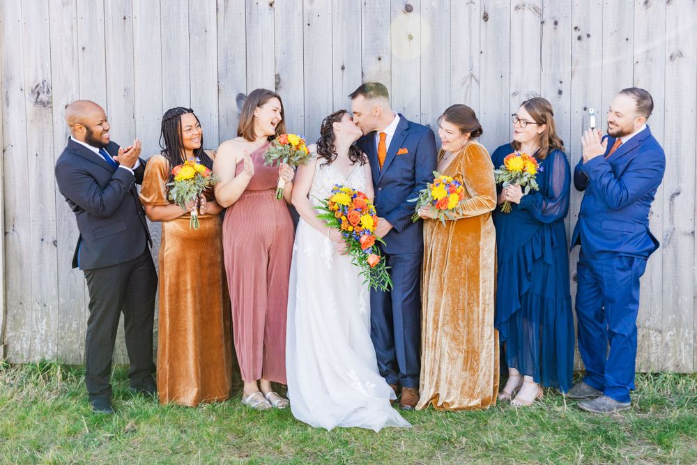 Joyful wedding party laughs together while posing against wooden fence backdrop.