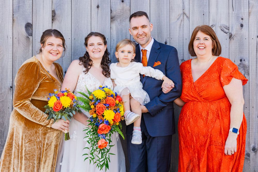Wedding group photo with colorful attire featuring orange and gold dresses against wooden backdrop.