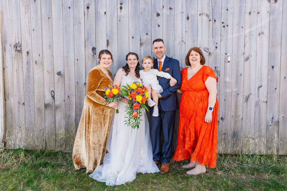 Family portrait with wedding party members in bright orange and gold dresses against rustic fence.