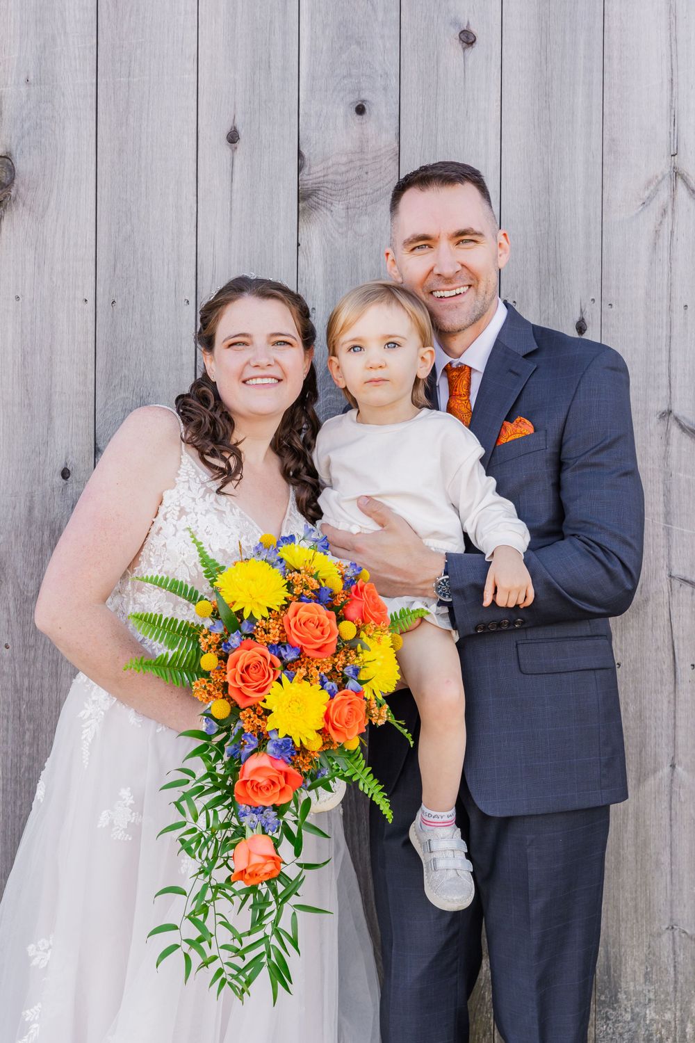 Wedding portrait against wooden fence with bright orange and yellow cascading bouquet.