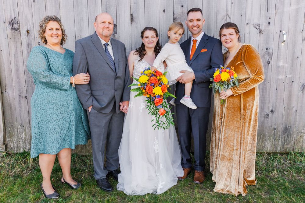 Multi-generational family wedding photo featuring formal attire and bright florals against wooden backdrop.