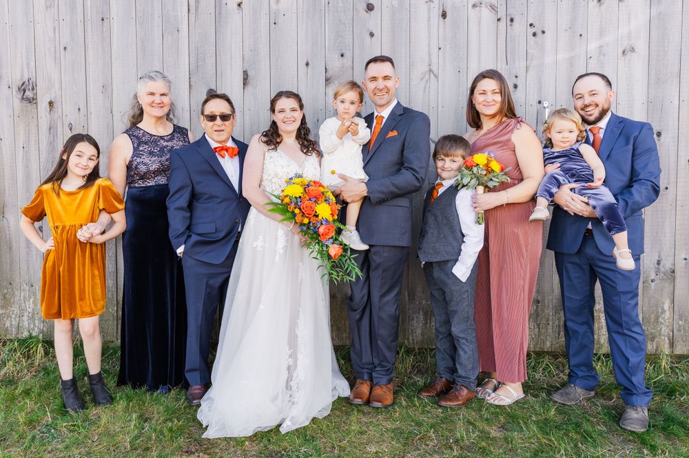 Extended family wedding portrait with children and adults in formal attire against rustic wooden wall.