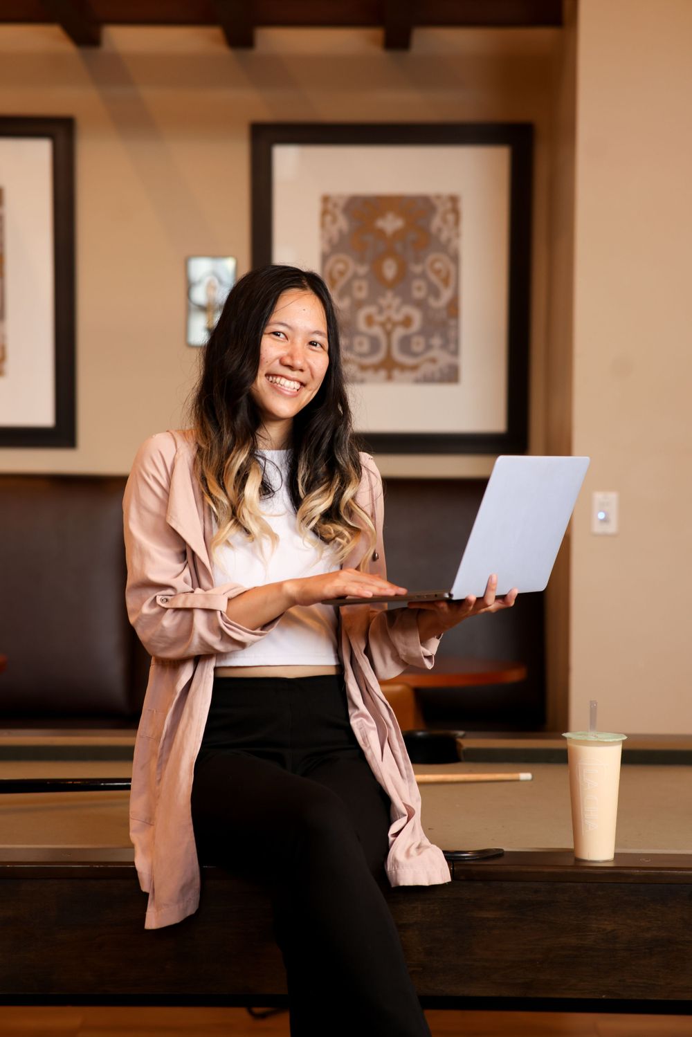 Professional wearing a pink cardigan holds papers while sitting casually in an office setting with framed artwork.
