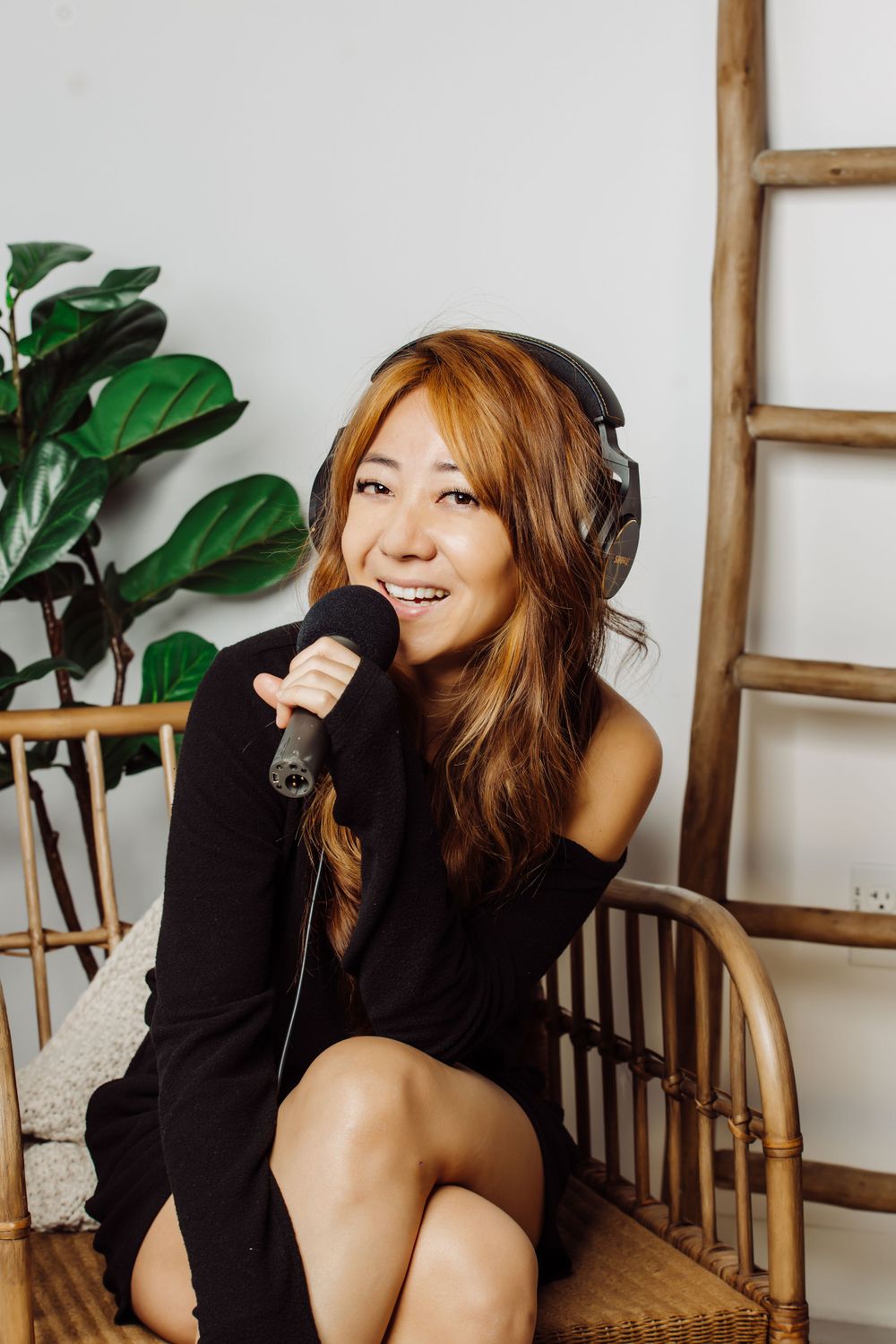 Singer in a black sweater performs with a microphone while sitting in a rattan chair next to indoor plants.