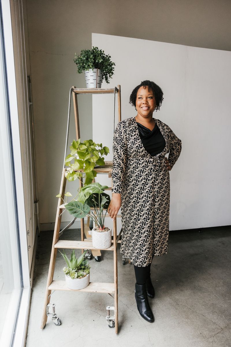 Professional wearing a leopard print dress poses with a decorative ladder shelf displaying potted plants near a window.