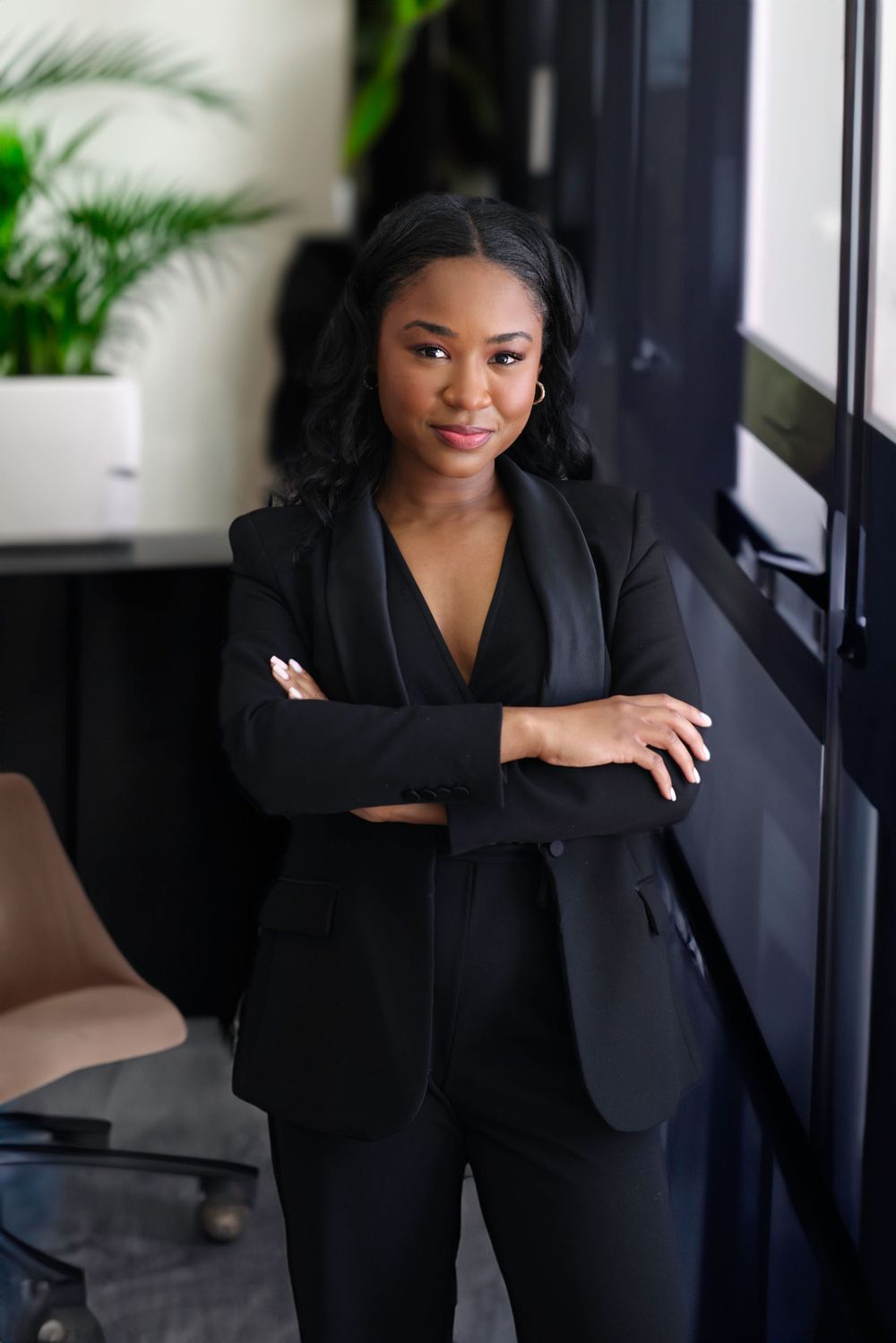 Professional in black business suit poses confidently near window with indoor plants.
