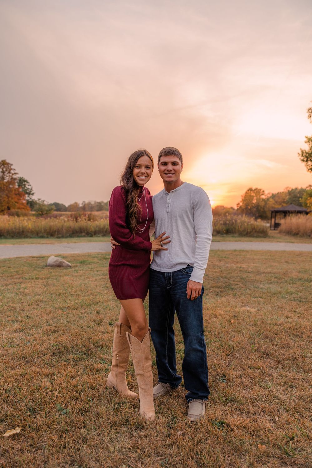 A couple in matching casual fall outfits stands arm in arm on a grassy lawn as the sun sets behind them.