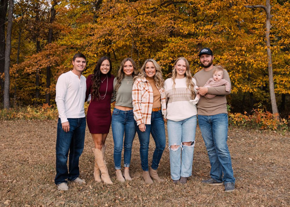 Family group with a baby poses in a row beneath vibrant orange autumn trees in a fall family portrait.